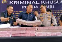 Direktur Reserse Kriminal Khusus (Dirreskrimsus) Polda Jambi, Kombes Pol Taufik Nurmandia (tengah), bersama Kabid Humas Polda Jambi, Kombes Pol Erlan Munaji (kanan), menunjukkan sebagian barang bukti uang tunai senilai Rp18,9 miliar yang berhasil disita dan dibekukan dari jaringan peretas internasional pembobol 6.609 rekening nasabah Bank Jambi, dalam konferensi pers di Mapolda Jambi, Selasa (14/7/2026). (FOTO : Dok. Kompas.com)