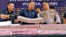 Direktur Reserse Kriminal Khusus (Dirreskrimsus) Polda Jambi, Kombes Pol Taufik Nurmandia (tengah), bersama Kabid Humas Polda Jambi, Kombes Pol Erlan Munaji (kanan), menunjukkan sebagian barang bukti uang tunai senilai Rp18,9 miliar yang berhasil disita dan dibekukan dari jaringan peretas internasional pembobol 6.609 rekening nasabah Bank Jambi, dalam konferensi pers di Mapolda Jambi, Selasa (14/7/2026). (FOTO : Dok. Kompas.com)